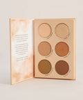 An open makeup palette with six circular eyeshadow pans in neutral and earthy tones, set against a clean light beige background.