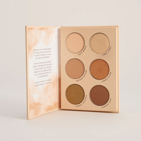 An open makeup palette with six circular eyeshadow pans in neutral and earthy tones, set against a clean light beige background.