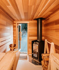 A cozy wooden sauna interior with light wood walls and benches, a black wood-burning stove with a tall chimney pipe, stacked firewood, and an open door revealing an outdoor view.