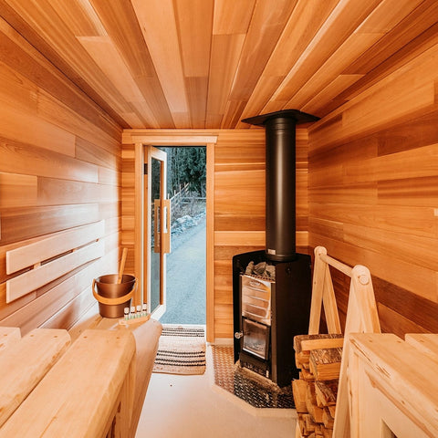 A cozy wooden sauna interior with light wood walls and benches, a black wood-burning stove with a tall chimney pipe, stacked firewood, and an open door revealing an outdoor view.