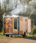 A small wooden sauna trailer with open doors and a visible wood-burning stove, shown outdoors on a paved surface with trees and smoke in the background.