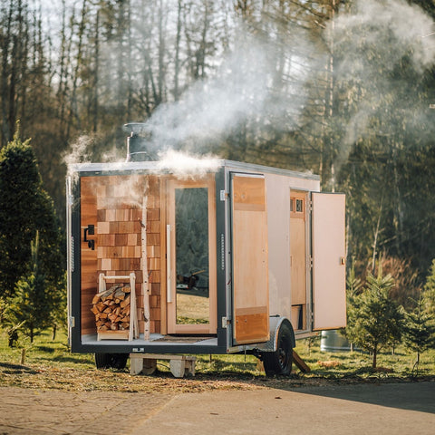 A small wooden sauna trailer with open doors and a visible wood-burning stove, shown outdoors on a paved surface with trees and smoke in the background.