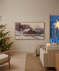 A framed winter landscape print hangs on a beige wall in a modern, cozy living room. The print depicts a snowy scene with a rustic cabin, bare trees, and a soft purple sky.