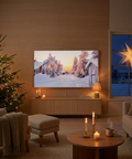 A large flat-screen TV mounted on a wood-paneled wall in a cozy living room, displaying a snowy winter village scene. The room includes a Christmas tree, armchair with throw blanket, ottoman, round coffee table with candles, sofa, and a window showing a glowing star outside.